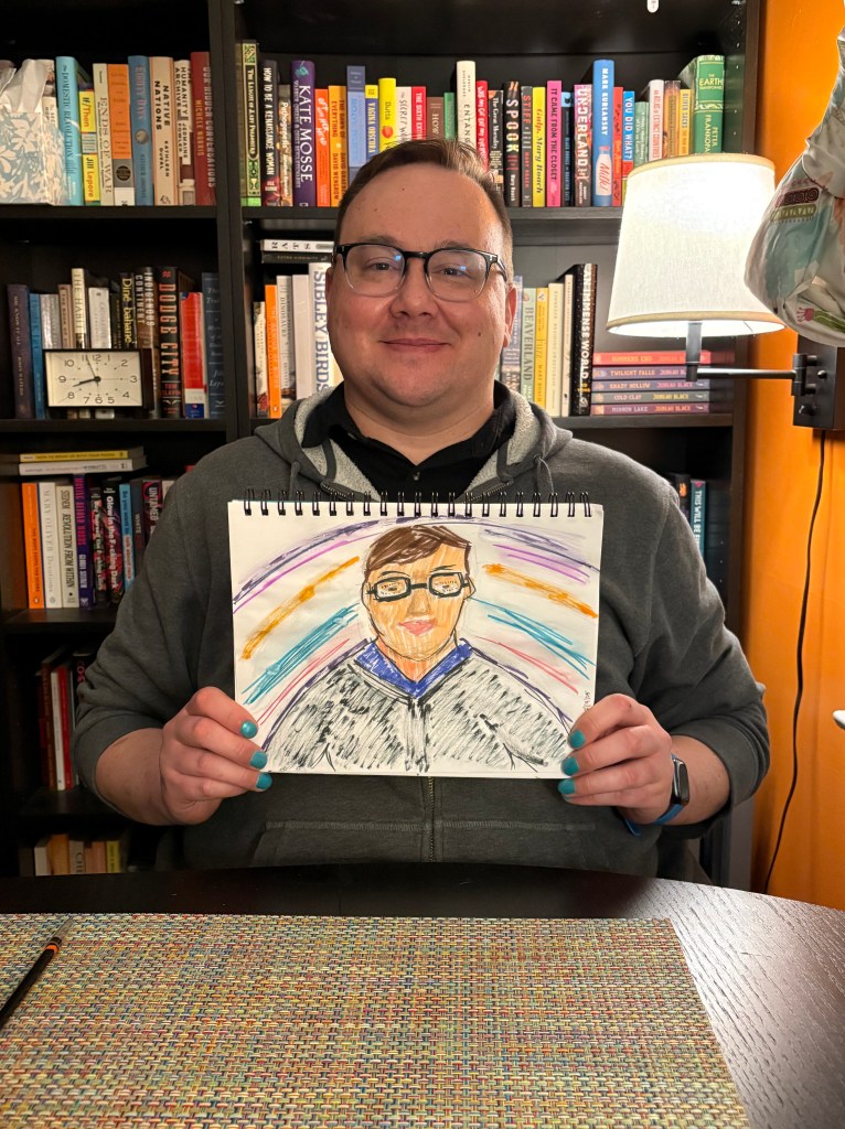 Josh sits in front of the bookcase in the dining room, and he is holding a painting of himself on a sketch pad.