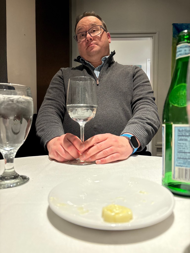 Josh holds a glass of sparkling water at a restaurant while sitting up straight to demonstrate that the table was way too short. 