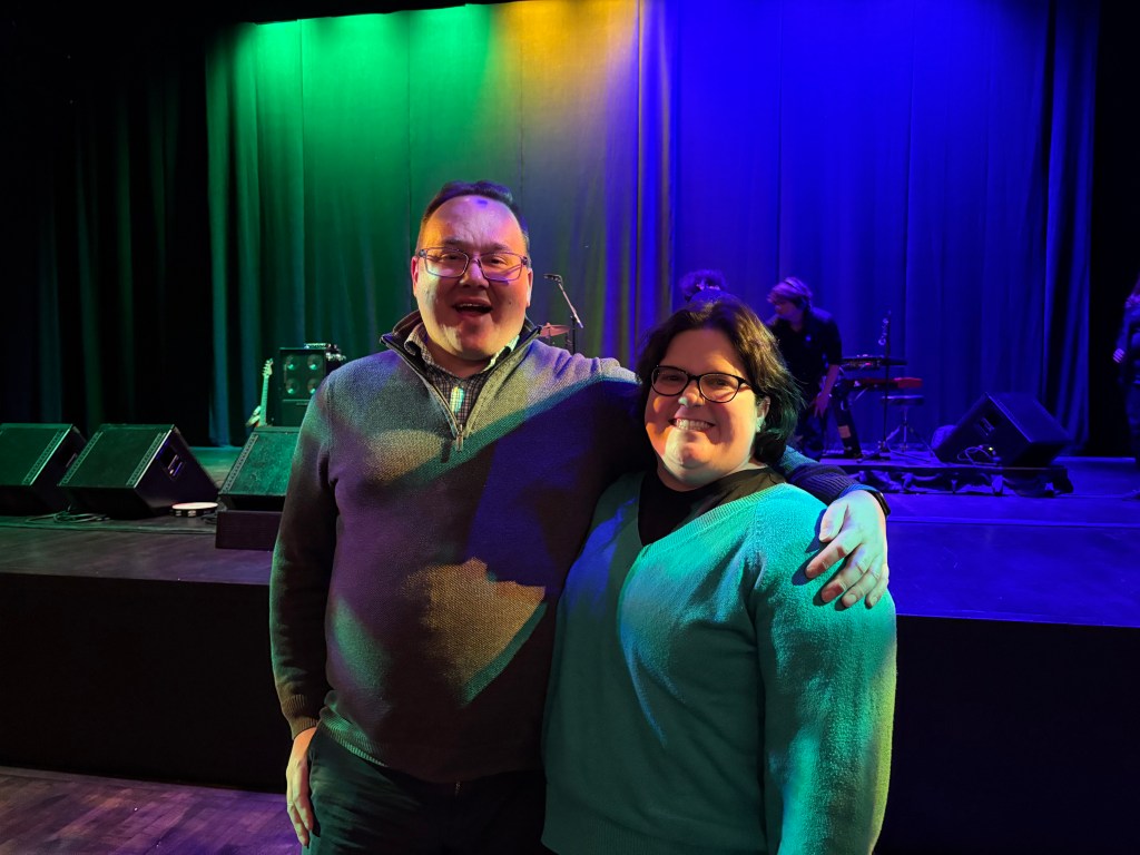 Josh and Jahnna smile in front of the stage at a concert venue in town. The curtain backdrop is lit up in Mardi Gras colors.